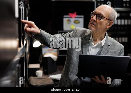 Senior private detective in criminal cases archive room looking for criminal case file details on cabinet shelves. Elderly investigator surrounded by criminology folders in agency depository office Stock Photo