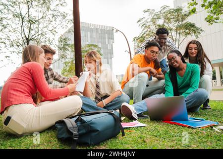 A happy group of young student people, sitting on the grass and sharing a laptop to do the homeworks, with smiles on their faces. Real multiracial teenage friends learning at university campus lawn. High quality photo Stock Photo