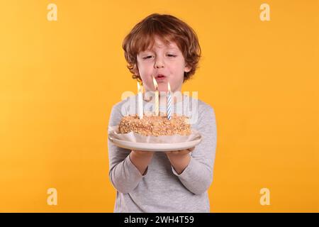 Birthday celebration. Cute little boy blowing candles on tasty cake against orange background Stock Photo