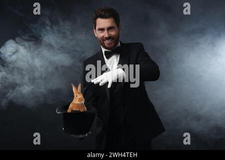 Magician showing trick with top hat and rabbit in smoke on dark background Stock Photo