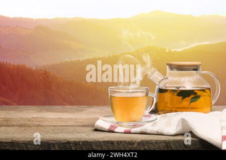 Aromatic green tea in glass cup and teapot on wooden table in mountains, space for text Stock Photo