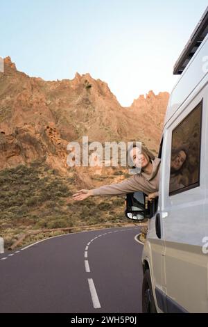 A man is joyfully extending her arm out of a white van, while traveling on a winding mountain road during the warm glow of sunset. Stock Photo