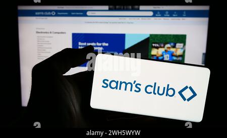 Person holding mobile phone with logo of American membership-only retail stores company Sam's Club in front of web page. Focus on phone display. Stock Photo
