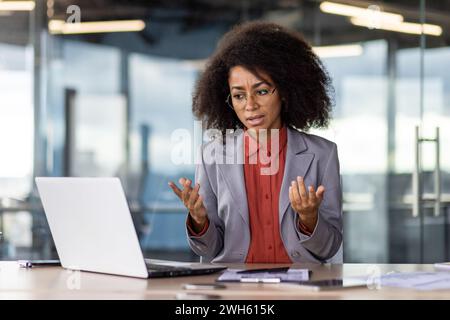 Professional young businesswoman with curly hair in office setting, showing concern while working on laptop at her desk. Stock Photo