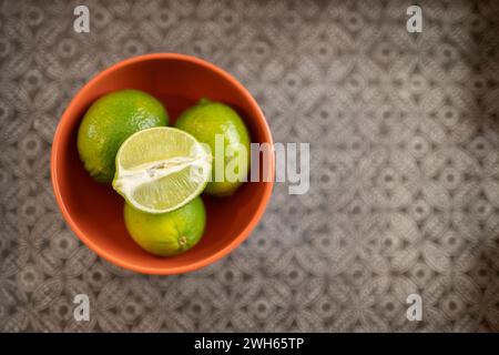 A top view of limes in an orange bowl on a silver-patterned background, empty right-hand side space Stock Photo