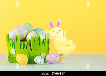 Happy Easter. Colored easter painted eggs in a green basket next to a chick with rabbit ears over yellow background Stock Photo