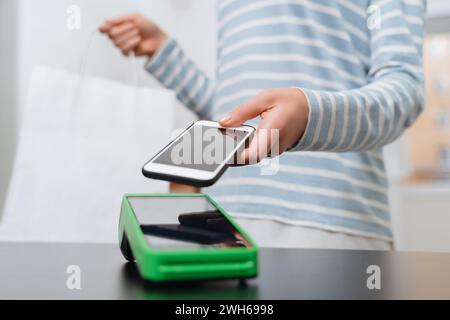 Female customer paying for purchase through smartphone using NFC technology. Closeup of woman's hand making payment through contactless machine or NFC terminal. Contactless payment concept Stock Photo