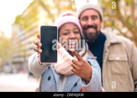 Happy multiracial young couple smiling pointing to empty smartphone screen outdoors Stock Photo