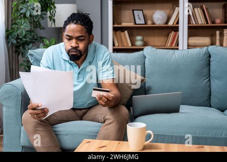 Focused serious African American businessman accountant analyst overspending holding documents looking at credit card laptop computer doing online tra Stock Photo