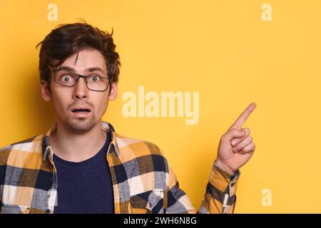 Portrait of confused man in glasses and casual shirt pointing on something with shocked face, isolated on yellow wall, copy space for text, advertisement. OMG, WOW, human reaction and emotions concept Stock Photo