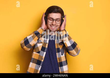 Delighted happy carefree man with eyeglasses listening to his favorite music with wireless headphones, dancing in rhythm of melody with closed eyes, has fun. Yellow background. Music concept Stock Photo