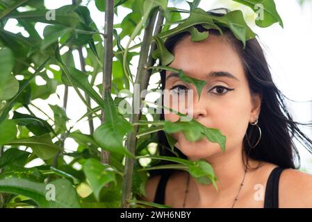 Face portrait of beautiful young woman with black hair looking at the camera through the leaves of a plant. Person traveling. Stock Photo