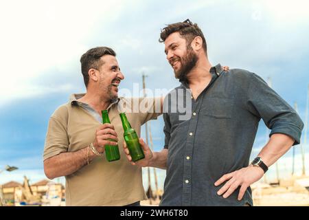 Two joyful men clinking beer bottles, celebrating friendship by the harbor. Stock Photo