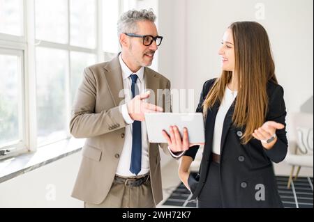 Grey-haired businessman engages with younger colleague, sharing a tablet screen in bright, modern office space. Their animated conversation and collaborative posture underscore concept of mentorship Stock Photo