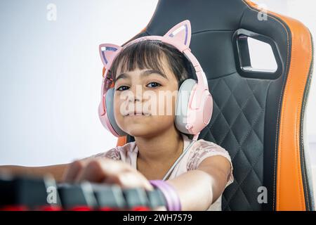 Portrait of a little girl with PC gamer accessories. She looks at the camera. She wears headphones with cat ears and is sitting in a gaming chair in f Stock Photo