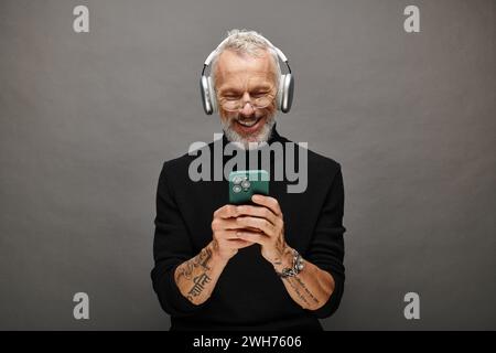 joyous attractive mature male model with beard and headphones in black turtleneck looking at phone Stock Photo