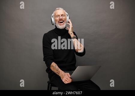 joyful handsome man with glasses and headphones sitting on chair with laptop and smiling at camera Stock Photo