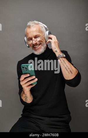 joyous attractive mature male model with beard and headphones in black turtleneck looking at phone Stock Photo