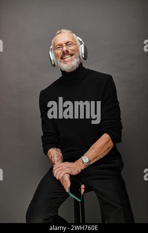 cheerful mature man in black turtleneck with headphones holding smartphone and looking at camera Stock Photo