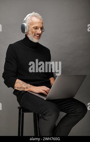 joyful handsome man with glasses and headphones sitting on chair with laptop and smiling sincerely Stock Photo
