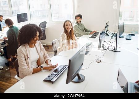 A team of professionals works in a brightly lit open space office. The diverse group is focused on their computer screens, suggesting a collaborative and tech-savvy workplace Stock Photo