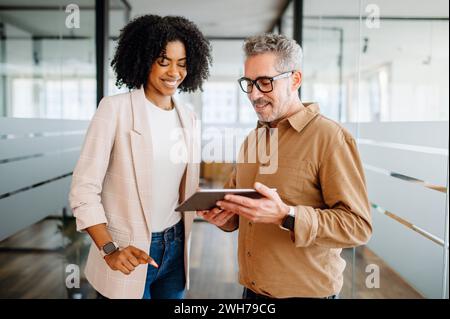 A businesswoman and a senior male colleague collaboratively review content on a tablet in a bright office space, reflecting a multigenerational workplace where experience meets new technology Stock Photo