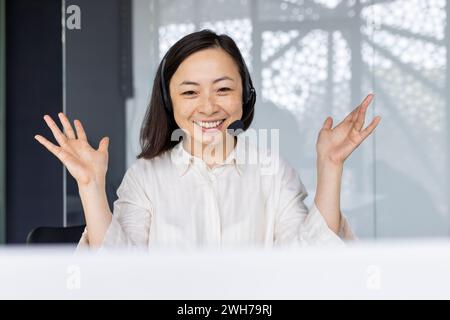 Professional and cheerful Asian female office worker wearing a headset, engaging with clients or team in a bright office space. Stock Photo
