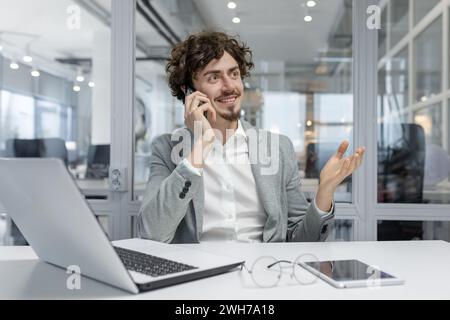 Curly-haired young professional man in a business setting, actively engaged in conversation on a phone call. Stock Photo