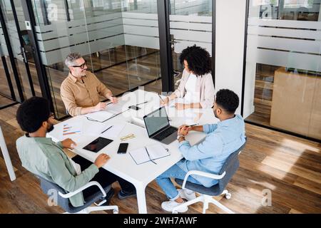 A team of four professionals are gathered around a white table in a modern office, indicative of a cooperative and strategic business meeting. Productive meetings and brainstorming concept Stock Photo
