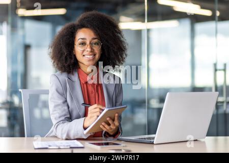A white-collar worker sits at a desk with a laptop and a tablet, surrounded by furniture. She smiles while working on her personal computer. Stock Photo