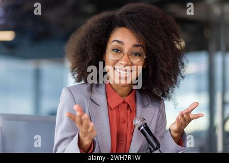 A happy woman, wearing a blazer, with a Jheri curl, smiles, and gestures while giving a speech at a music artist event, holding a microphone. Stock Photo