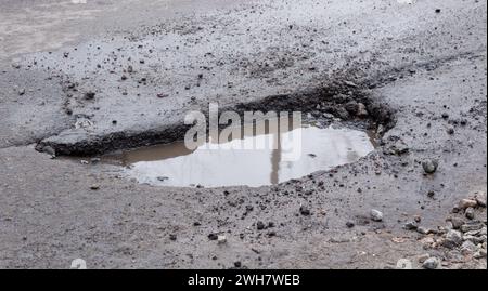 Deep pit on the road with puddle. Damaged asphalt after winter. Road repair. Stock Photo