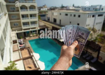 Man holds hundred dollar bills in front of a hotel with a swimming pool. Stock Photo