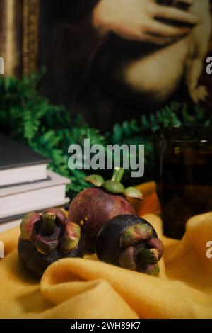 Lush still-life set up with three purple Mangosteens, books, vines, yellow felt cloth, a painting, glass of water. Close-up of sweet tropical fruit. Stock Photo