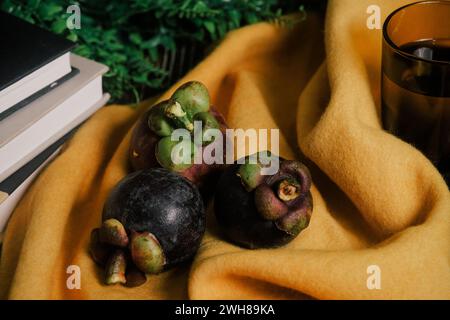 Close-up of three Mangosteens, a sweet tropical fruit. Lush still-life setup with vibrant yellow cloth, glass of water, stack of books, green vines. Stock Photo