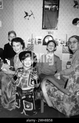 England. 1950s – A photograph depicting a woman, an elderly lady and three young boys in a domestic setting. The boy in the foreground is dressed as a cowboy, holding a pair of toy cap guns. In the background is a tiled fireplace, on which is placed a clock, greetings cards, candles and various ornaments, including a toby jug.  On the wall is a mirror and ceramic flying duck plaques. Stock Photo