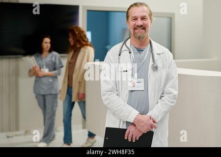 Successful male clinician in labcoat looking at camera with smile while standing against reception counter and two young women Stock Photo