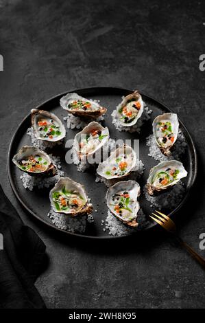 Freshly shucked oysters with buttermilk dressing, salmon roe, balsamic pearls and herb oil sitting on rock salt on a black plate Stock Photo