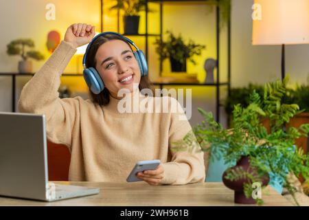 Happy relaxed overjoyed Caucasian young woman in wireless blue headphones sits at home table choosing listening favorite disco music in smartphone dancing entertaining. People weekend activities Stock Photo