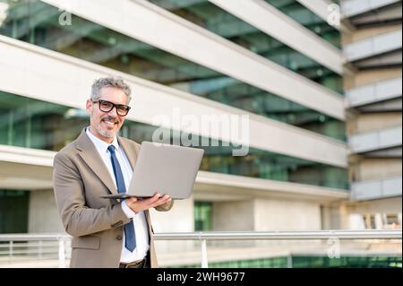 A confident senior 60s businessman in a brown suit stands outdoors, holding his laptop with an engaging smile, showcasing a fusion of experience and modern technology Stock Photo