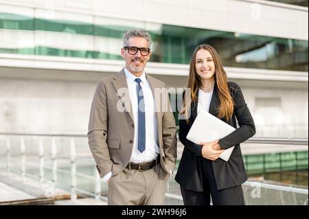 Standing side by side, the seasoned businessman and his young female colleague share a professional moment, woman holding laptops and both dressed in business attire against a modern building backdrop Stock Photo