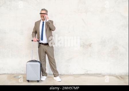 A happy senior businessman converses on his mobile phone while standing with his suitcase, demonstrating the constant connectivity of mobile professionals Stock Photo