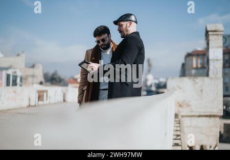 Two fashionable male professionals engage in a business discussion using a digital tablet, standing against an urban cityscape. Stock Photo