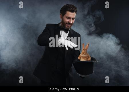 Magician showing trick with top hat and rabbit in smoke on dark background Stock Photo