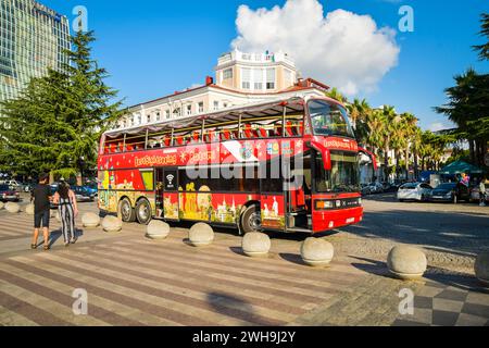 Batumi, Georgia - 4th august, 2022: female tourist walk us phone in street pass red two floor double-decker classic sightseeing bus around city. Famou Stock Photo