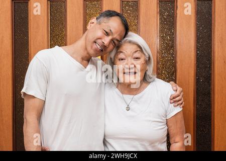 Happy older Asian mother and adult son hugging affectionately, looking at camera, smiling, enjoying family visit, free time. Stock Photo