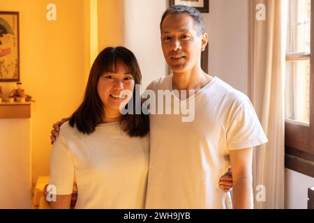 Adult siblings standing hugging each other of Japanese ethnicity, smiling and looking at the camera. Horizontal portrait indoors with warm light. Stock Photo