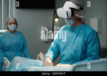 Mature surgeon in VR headset, scrubs, protective mask and gloves using medical equipment while performing surgical operation Stock Photo