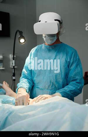 Mature male clinician in VR headset, medical scrubs, mask and gloves using operating equipment during surgical operation Stock Photo