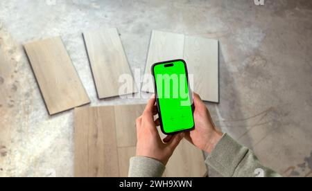 Hands Holding Device with Green Chroma Key Screen above Luxury Vinyl Wood Samples, Laminate On Floor In House. Choosing, Selecting Waterproof Flooring Stock Photo